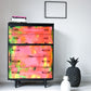 Colorful painted dresser with a vibrant pink, orange, and green abstract design, placed in a modern minimalist room with decorative pineapple sculptures, a black picture frame, and a white wall background