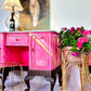 Vivid pink painted vintage sideboard decorated with vibrant pink and purple petunias in a wicker plant stand, featuring a painting with a pregnant woman on the wall, warm table lamp, and decorative items, creating a colorful and artistic home decor setting